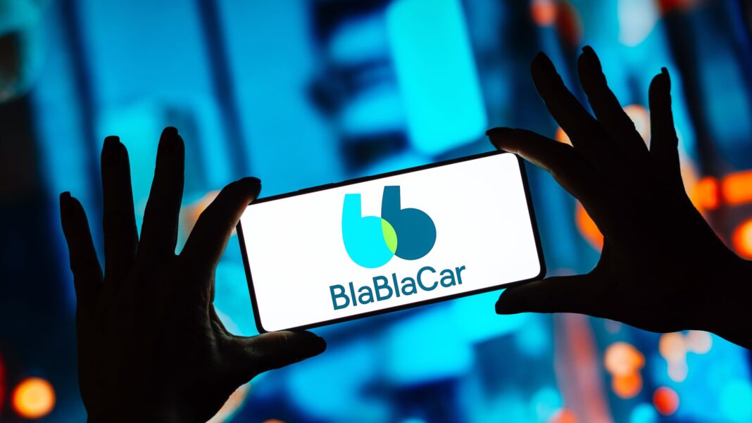 Людина тримає в руках смартфон із відкритим логотипом та назвою сервісу пошуку попутників BlaBlaCar на екрані на розмитому синьому тлі.
